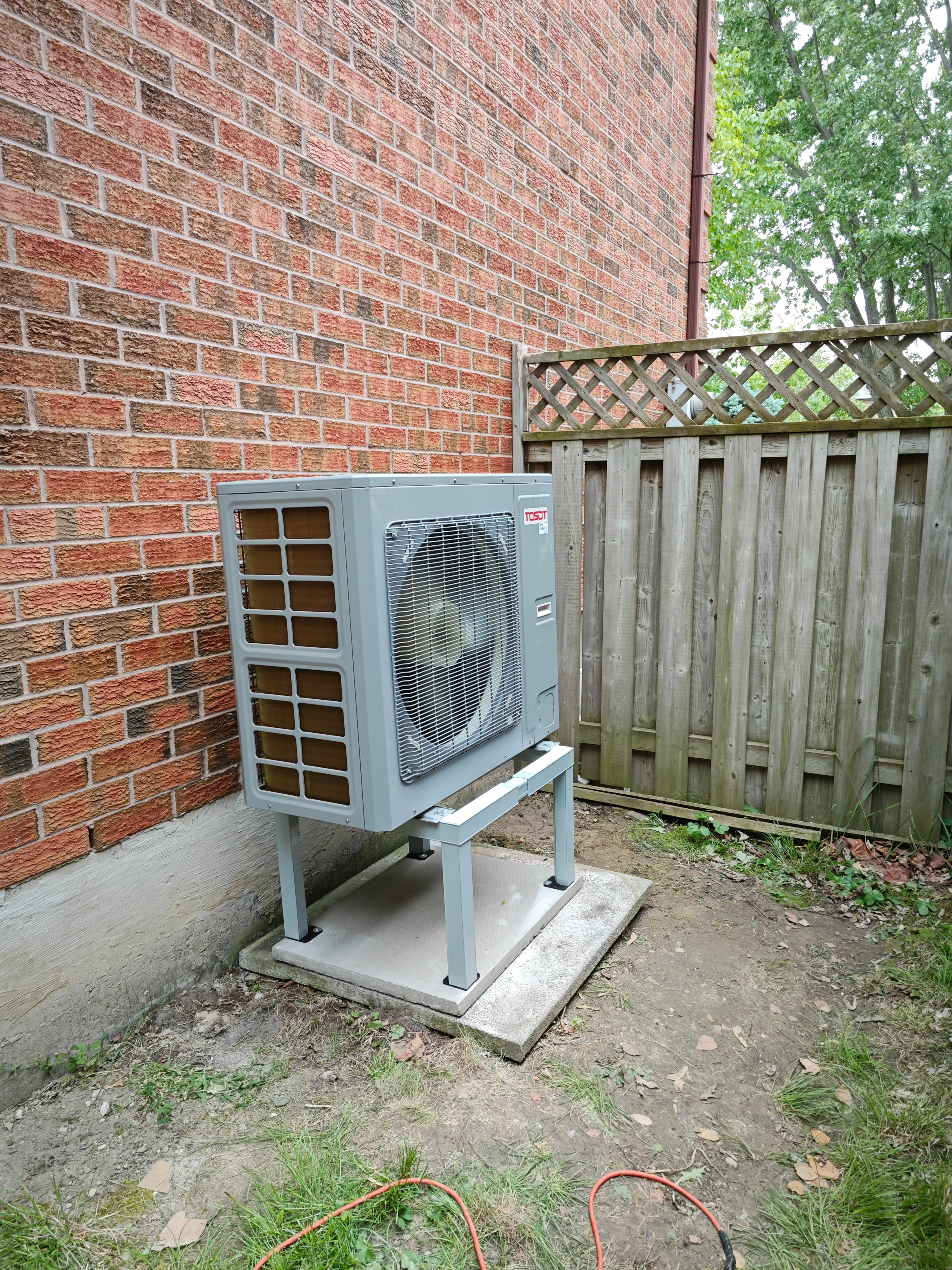 Gray outdoor heat pump on a stand beside a brick wall and a wooden fence